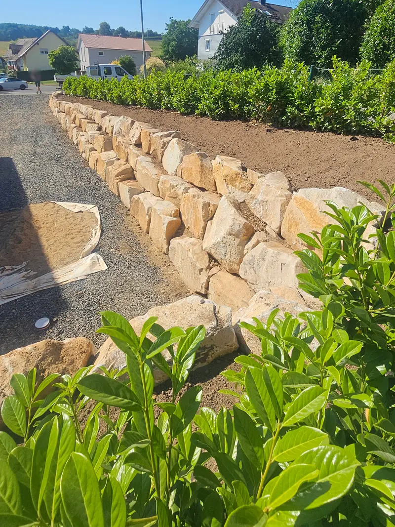 Mauer aus hellen Natursteinblöcken entlang der Zufahrt, darüber eine Hecke