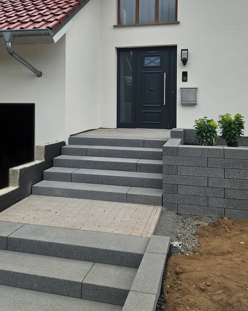 Außentreppe aus dunkelgrauen Blockstufen zur Haustür, rechts eine Mauer aus Mauersteinen