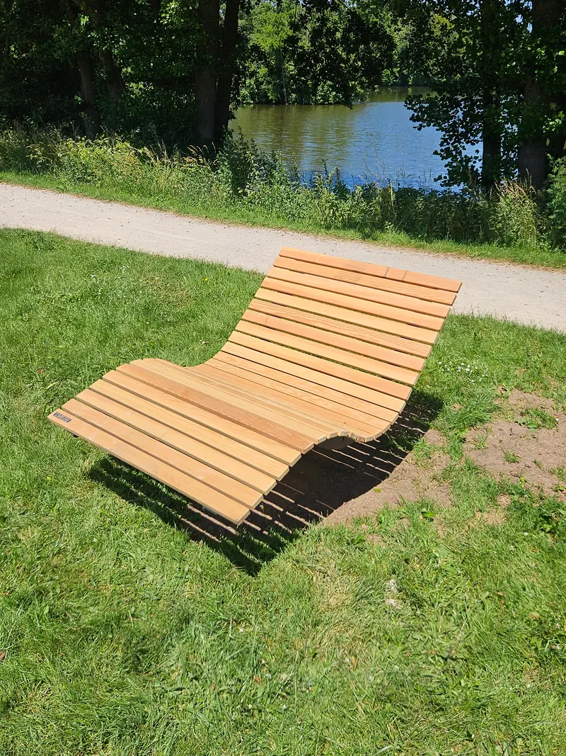 Liege aus Holz auf der Wiese am Weg, dahinter der Weiher