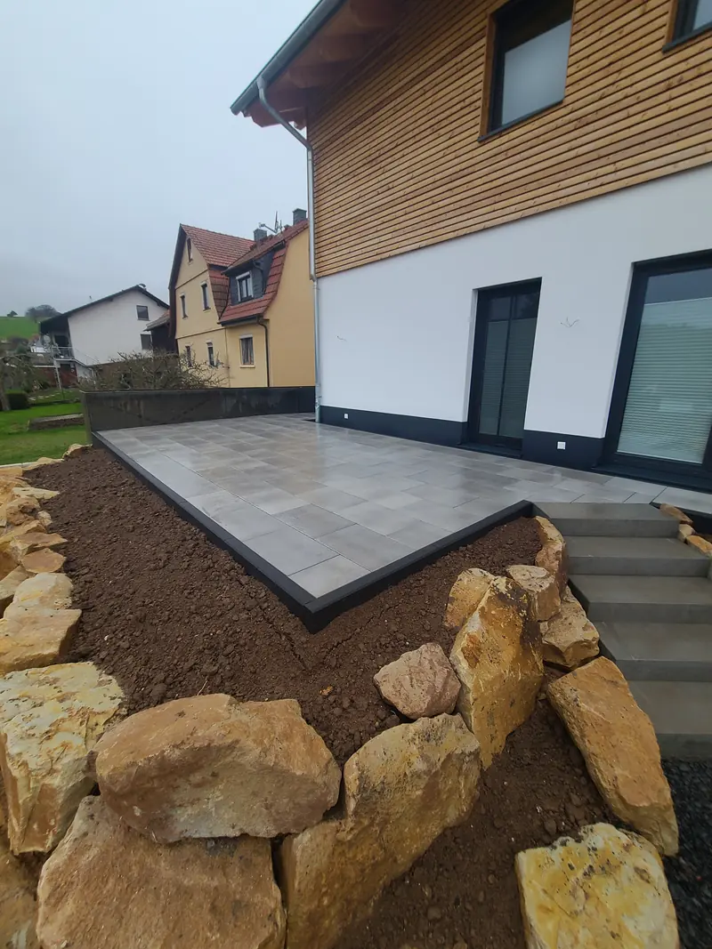 Terrasse aus großen Platten an einem Haus mit Holzfassade, davor Natursteine