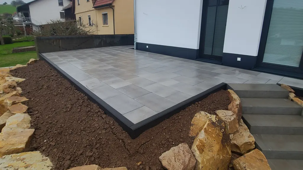 Terrasse aus großen grauen Platten mit dunkler Einfassung, davor Natursteine am Hang