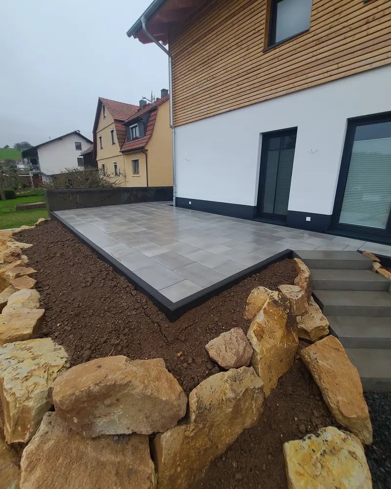 Terrasse aus großen grauen Platten mit dunkler Einfassung, davor Natursteine am Hang